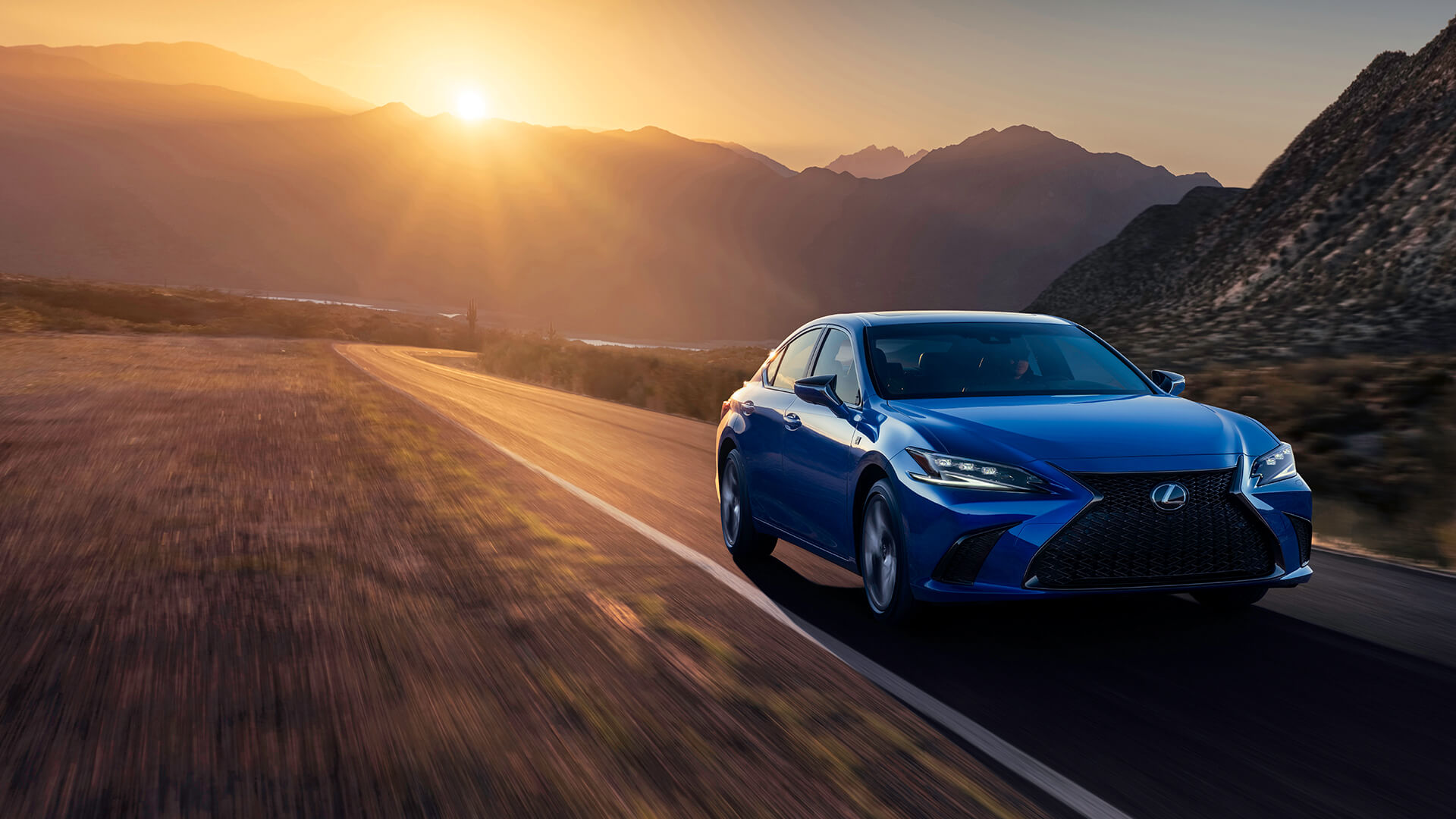 Blue Lexus ES is being driven on the road with a mountainous backdrop.