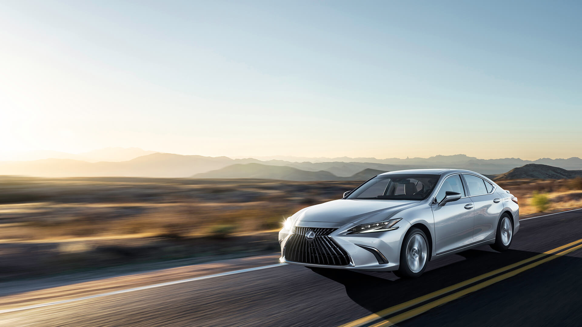 Silver Lexus ES is being driven on the road with a mountainous backdrop.