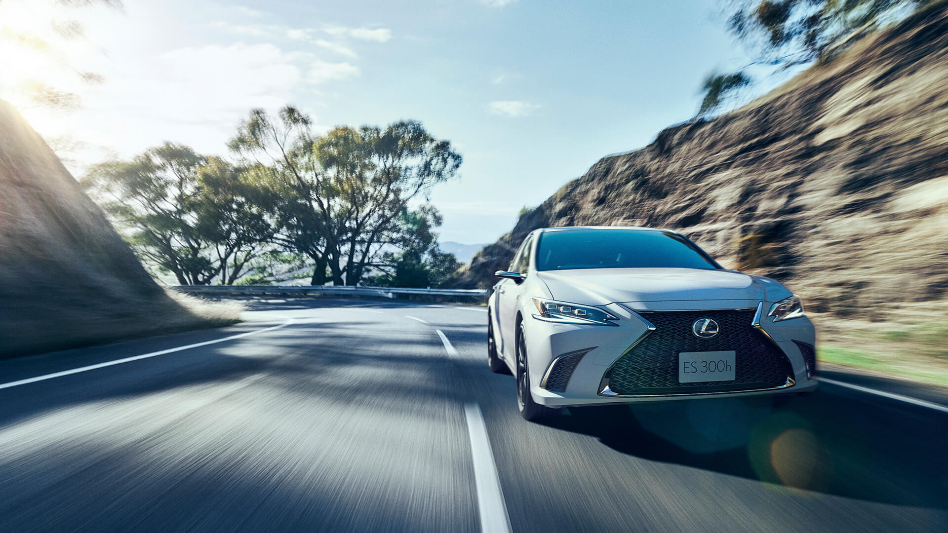Silver Lexus ES F SPORT is being driven on the road with a hilly backdrop.